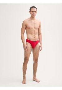 Maillot de bain rouge en tissu extensible, avec un design taille basse et un petit logo sur la ceinture. Texture lisse et coupe ajustée.