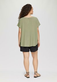 Olivgrünes, locker sitzendes Oberteil mit kurzen Ärmeln, geradem Saum und Schlüsselholedetail im Rücken, kombiniert mit schwarzen Shorts und Riemchensandalen.