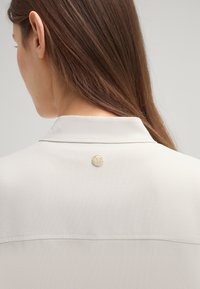 Lichtgrijze blouse met een gladde stofstructuur. Bevat een knoop aan de achterkant met een boomlogo en een gevouwen kraagontwerp.