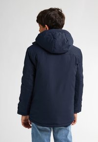 Cappotto imbottito blu navy con cappuccio rimovibile, maniche lunghe e vestibilità ampia, abbinato a jeans di denim blu chiaro.