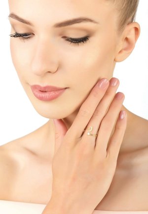 Gold double band ring featuring a zigzag design and a small clear stone accent. Model wearing it while touching her face.