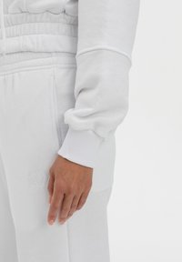 Felpa bianca con polsini a coste, dotata di cinturino elastico in vita. Il tessuto sembra morbido e testurizzato, con dettagli del logo ricamati.