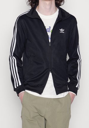 Personne portant une veste de survêtement Adidas noire zippée avec des bandes blanches sur les manches et un t-shirt blanc à motif en dessous, mains dans les poches du pantalon.
