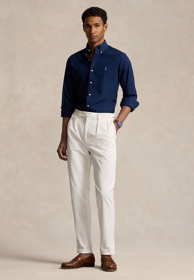 Mann steht mit Händen in den Taschen, trägt ein marineblaues Hemd mit Knöpfen, weiße Hose, braune Lederschuhe und eine Armbanduhr.