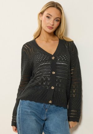 Blonde vrouw die een zwart opengewerkt vest met knopen en een blauwe spijkerbroek draagt, staand tegen een effen lichte muur.