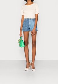 Top beige chiaro a maniche corte, pantaloni corti in denim blu a vita alta, scarpe con tacco trasparente e borsa verde con dettagli in oro.