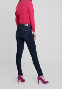 Jeans ajustados azul oscuro con bolsillos traseros y herrajes plateados, combinados con un suéter rosa brillante y tacones altos morados.