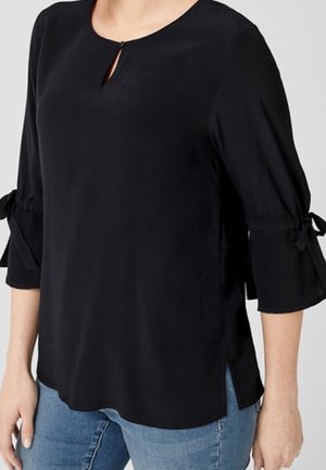 Kvinde iført en sort, langærmet bluse med bundne sløjfer på ærmerne og en lille forreste nøglehulsknap, kombineret med blå jeans.