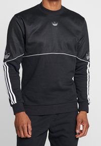 Sudadera negra de Adidas con ribetes blancos y logotipos de trébol en el pecho y las mangas, con detalles de tres rayas blancas en las mangas.