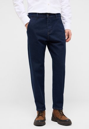 Mann trägt dunkelblaue Jeans, braune Schnürstiefel und ein weißes Langarmshirt mit einer Hand in der Tasche.