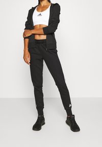 Tenue de sport noire composée d'un sweat à capuche zippé et de joggers ajustés, arborant le logo Adidas blancs et des bandes sur les côtés, assortie à des baskets noires.