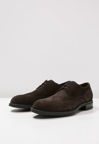 Chaussures derby en daim marron avec un bout arrondi, des détails brogues sur le cou-de-pied, et une semelle extérieure noire contrastante. Équipées de trois lacets.