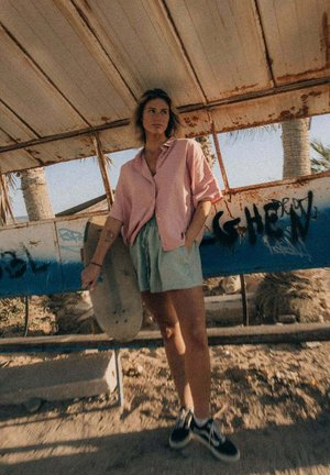 Młoda osoba w różowej koszulce i zielonych szortach stoi trzymając deskorolkę pod zardzewiałym metalowym schronieniem z graffiti i palmami.