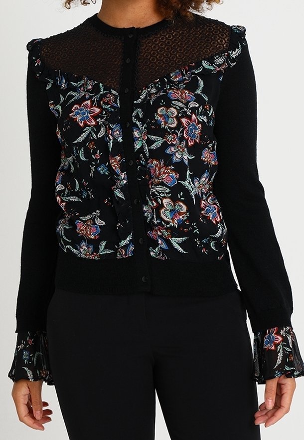 Cardigan noir à manches longues avec motif floral, empiècement en dentelle transparente, détails froufroutants sur le devant et aux poignets, fermeture boutonnée, porté avec un pantalon noir.