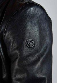 Veste en cuir noire avec une texture lisse, un design ajusté et un détail de logo noir circulaire sur l'épaule gauche.
