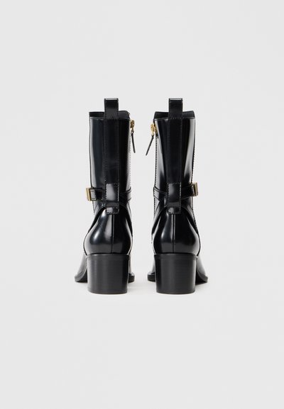 Un par de botas de cuero negras hasta la mitad de la pantorrilla con tacones cuadrados, cremalleras laterales y correas decorativas con hebilla, vistas desde la parte posterior sobre un fondo liso.
