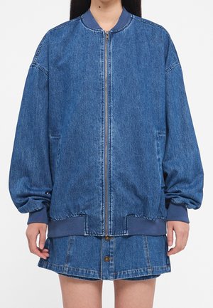 Personne portant une veste en jean bleu zippée avec des poignets et un col côtelés, associée à une jupe en jean assortie à boutons sur le devant, sur fond blanc.