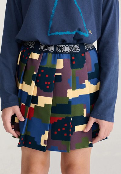 Enfant portant une jupe à motifs géométriques colorés avec des plis et un haut bleu marine avec un triangle souligné en bleu.