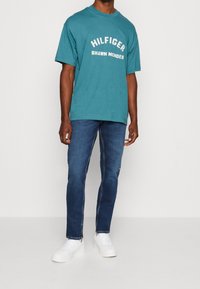 Turkusowa koszulka z krótkim rękawem z białym napisem "HILFIGER" oraz "SHAWN MENDES", zestawiona z niebieskimi dżinsami i białymi trampkami. Prosty, casualowy strój.