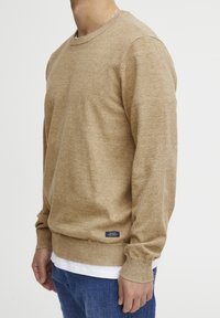 Beige gebreide trui met een ronde halslijn, geribde manchetten en zoom, met een klein blauw label op de taille. Draag over een wit t-shirt.
