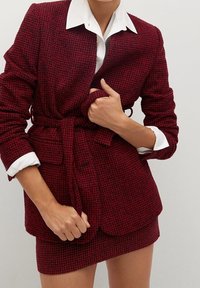 Nainen säätää vyöllä varustettua punamusta houndstooth-takkia, joka on puettu yhteensopivan hameen ja valkoisen kauluspainon, jonka hihansuut on käännetty ylöspäin, päälle.