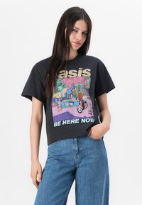 T-shirt nera in cotone con un design grafico colorato che presenta il testo "Oasis" e "Be Here Now" in colori a contrasto, abbinata a jeans blu.