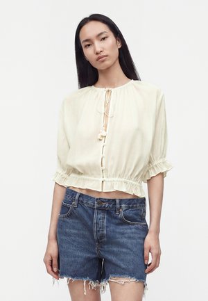 Femme portant une blouse crème à manches courtes avec des liens et un short en jean bleu effiloché, debout devant un fond blanc uni.