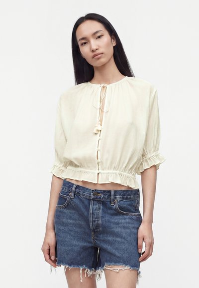Femme portant une blouse crème à manches courtes avec des liens et un short en jean bleu effiloché, debout devant un fond blanc uni.