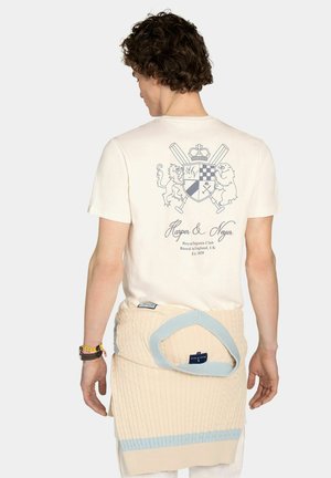 Joven con cabello rizado que lleva una camiseta blanca con un escudo real y texto en la parte trasera, y un suéter de punto grueso color crema atado alrededor de la cintura.