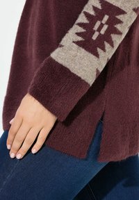 Burgunderfarbener Pullover aus weichem, strukturiertem Stoff mit einem beigen gemusterten Ärmel und seitlichen Schlitzen. Kombiniert mit dunkelblauen Jeans.