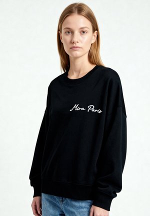 Młoda kobieta o prostych blond włosach nosi czarną bluzę z haftowanym napisem "Mira Paris" oraz jasnoniebieskie dżinsy na jednolitym tle.