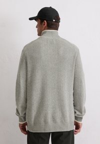 Pull en maille côtelée gris clair avec col montant et épaules tombantes. Présente un tricot texturé ainsi qu'une taille et des poignets à contraste.