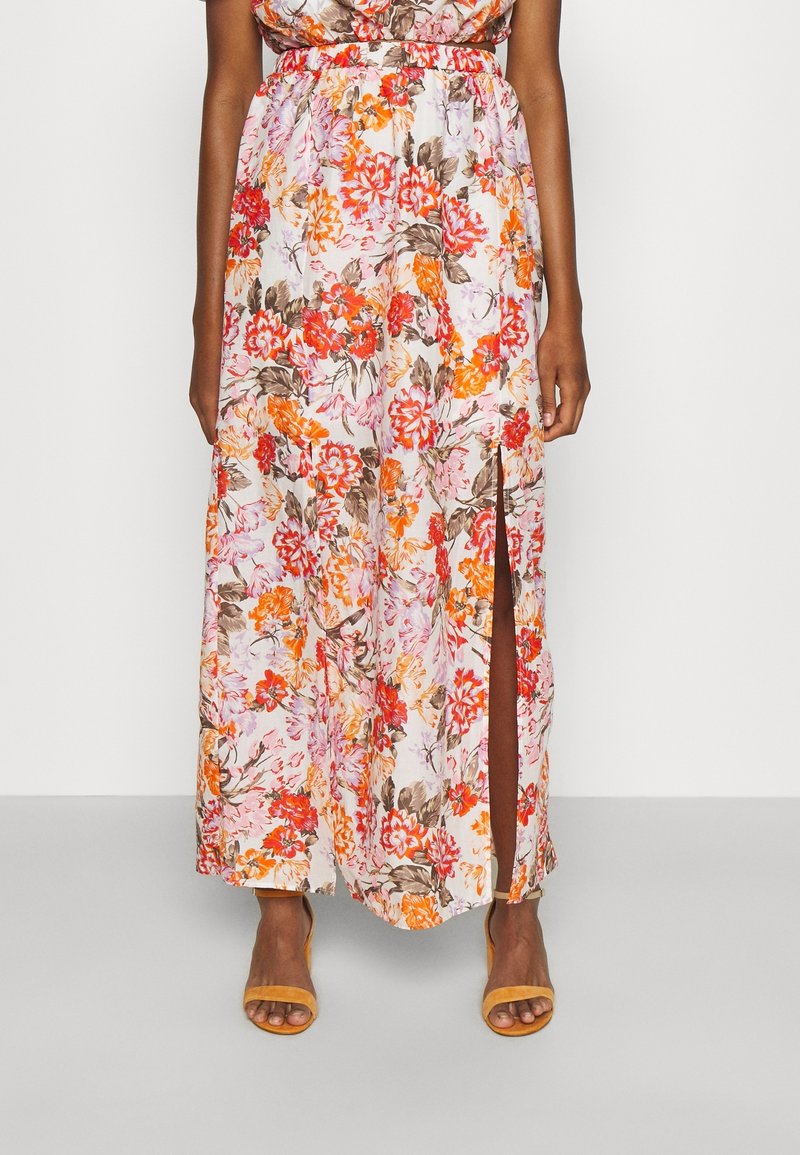 Femme portant une jupe longue blanche avec un imprimé floral orange, rouge et marron, fendue sur le côté, associée à des talons ouverts jaunes.