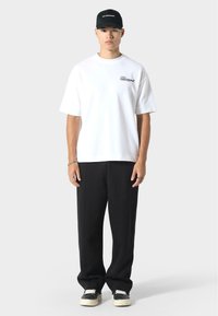 Biała, oversize'owa koszulka z czarnym logo, zestawiona z czarnymi, luźnymi spodniami i czarnymi sneakersami z białą podeszwą. Całość dopełnia czarna czapka.
