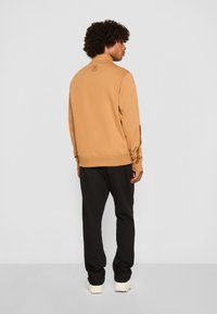 Tan sweatshirt med høj hætte og logo-patch på ryggen, parret med sorte bukser; modelen står med ryggen til mod en ensfarvet baggrund.