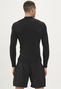 Svart långärmad träningströja med strukturerad design, figurnära passform och mesh-detaljer vid axlarna, ihopparad med svarta shorts.