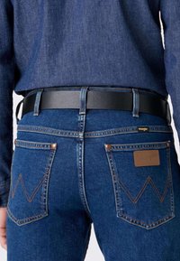Blå denim jeans med høj talje, sort læderbælte, gyldne syninger og karakteristiske baglomme-designs med en 'W' form.