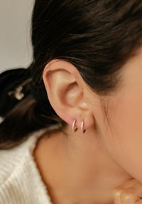 Boucles d'oreilles créoles dorées avec de petites pierres claires disposées sur le devant, présentées sur l'oreille d'un modèle. La texture semble lisse avec une finition métallique polie.