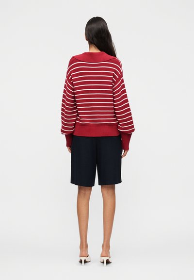 Γυναίκα με μακριά σκούρα μαλλιά που φοράει κόκκινο και άσπρο ριγέ πουλόβερ, μαύρες σορτς μέχρι το γόνατο και λευκά mules με τακούνι, όρθια και στραμμένη προς τα πίσω.