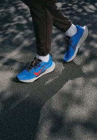 Chaussures de sport bleues avec une tige texturée et des accents rouges contrastants, dotées d'une semelle épaisse blanche et de lacets marron, portées au pied d'une personne.