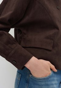 Braune Wildlederjacke mit umgeschlagenem Kragen und Knöpfen, kombiniert mit blauen Denim-Jeans. Der Stoff hat eine weiche Textur.