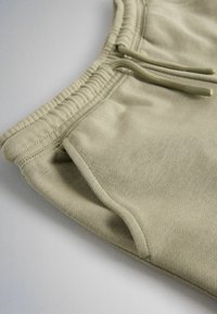 Détail d'un pantalon de jogging beige montrant une taille élastique avec cordons de serrage et une poche latérale sur un fond blanc lisse.