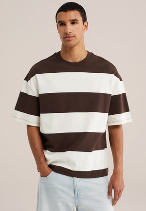 Ung mand med kort mørkt hår iført en brun og hvid bredstribet kortærmet skjorte og lyseblå jeans med den ene hånd i lommen.