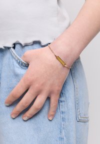 Złota bransoletka z prostokątnym zawieszką, przymocowana do łańcuszka, noszona na ręce opierającej się na jasnoniebieskim dżinsowym materiale. Prosty i elegancki design.