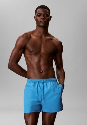 Athletischer Mann in leuchtend blauen Badeshorts, der mit einer Hand in der Tasche vor einem hellgrauen Hintergrund steht.