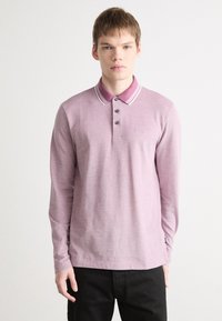 Polo majica s dugim rukavima u svijetlo ružičastoj boji, s kontrastnim ovratnikom sa prugama, pet dugmadi i malim logom na prsima.