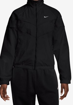Osoba nosząca czarną kurtkę Nike na zamek i pasujące czarne spodnie z logo Nike na piersi i udzie.