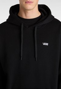 Felpa nera con cappuccio in cotone, dotata di tasca frontale, lacci e un piccolo logo bianco "VANS" sul petto. Texture morbida.