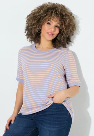 Femme aux cheveux bouclés portant un t-shirt rayé lilas et pêche et un jean bleu, debout devant un fond blanc avec une main dans la poche.