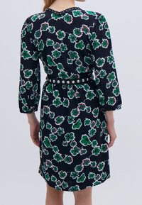 Vestido floral con un fondo negro y patrones de flores verdes y blancas, mangas tres cuartos y un cinturón en la cintura con clavos plateados.